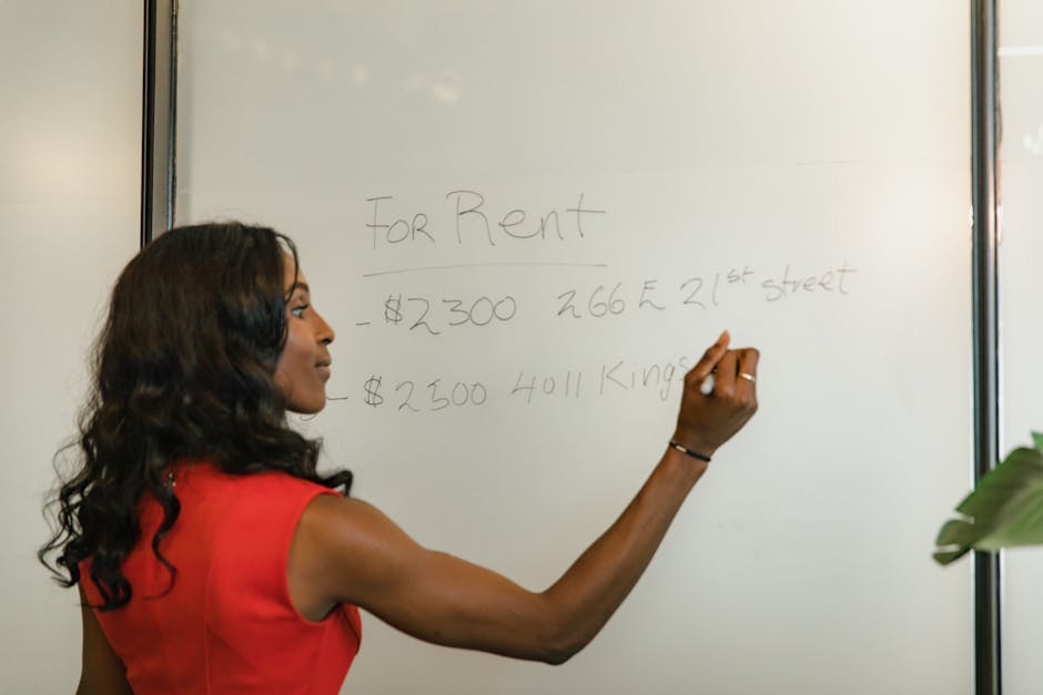 A woman with long, wavy black hair and wearing a sleeveless red top is standing in front of a whiteboard, writing rental prices and addresses with a black marker. The whiteboard shows handwritten text including 'For Rent,' a price of '$2300,' an address '266 E 21st Street,' and another rent price of '$2500' for '4011 Kings.' The room appears to be well-lit, with part of a green plant visible in the lower right corner. The setting suggests an office or meeting room, emphasizing a clean and organized environment suitable for professional discussions, with the surfaces in the room appearing tidy and dust-free. The presence of the whiteboard and the woman's focused activity highlight a setting related to real estate or property management, aligning with the context of surface and document cleaning, similar to the services offered by Carpet Cleaning SE10 for maintaining hygiene and cleanliness in commercial spaces.