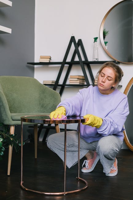 A woman with blonde hair tied back, dressed in a purple sweatshirt and grey checked pants, is kneeling on the dark wooden floor of a living room, wearing yellow rubber gloves. She is carefully cleaning a small, round, glass-topped table with a pink cloth, appearing focused on her task. In the background, there is a green upholstered armchair, a black leaning shelf holding small stacked books, a decorative plant, and a spray bottle, along with a large, round wall mirror reflecting part of the room. The room is well-lit with natural light, and the overall scene emphasizes thorough surface cleaning and maintenance, with attention to hygiene in a modern domestic setting. This environment is associated with professional carpet and upholstery cleaning services, as promoted by Carpet Cleaning SE10.