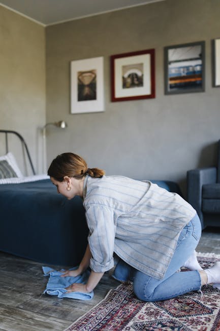 A professional cleaner dressed in a white protective suit, face mask, and gloves is performing deep carpet cleaning in a bright, modern living room. The cleaner uses a handheld steam cleaning machine
