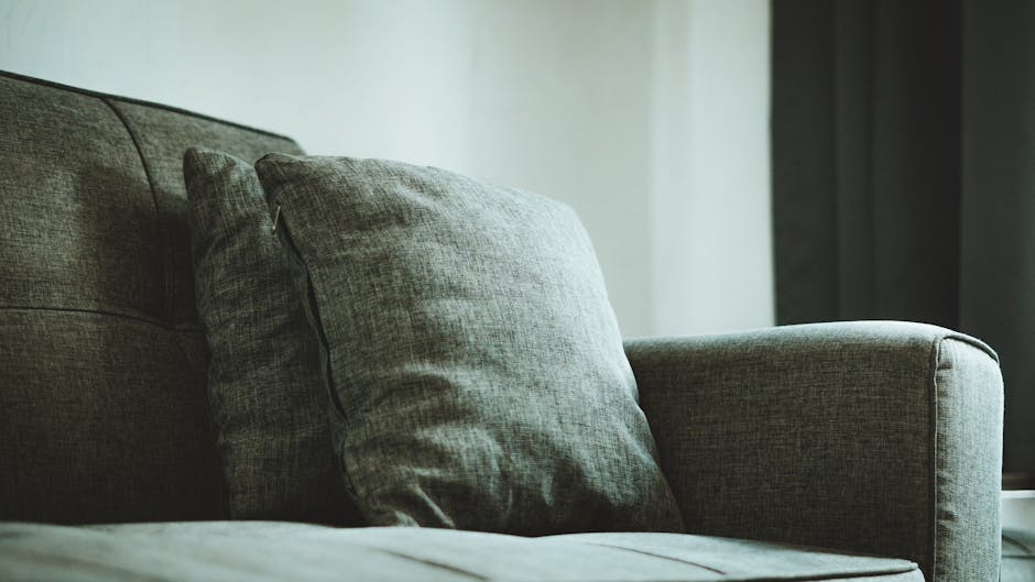 Close-up of a grey upholstered sofa in a living room setting, featuring a soft fabric surface with two matching cushions neatly arranged. The sofa appears clean and well-maintained, with minimal visible dust or stains. The room has natural lighting coming from the right, illuminating the furniture and creating a calm, tidy atmosphere. The background includes a plain, light-colored wall and a partially visible dark curtain, indicating a tidy, modern domestic space. The image reflects professional upholstery cleaning and surface cleaning practices, highlighting the importance of regular maintenance for a hygienic and visually appealing environment, as provided by Carpet Cleaning SE10.