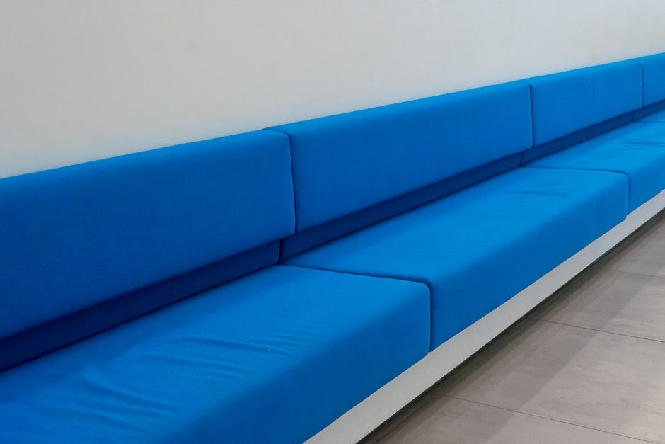 A long, modern blue upholstered bench with clean and smooth cushions, situated along a white wall in a well-lit room. The bench has a sleek design with consistent padding and is positioned on a light gray tiled floor. The upholstery appears pristine and free of stains, reflecting a high level of surface cleaning and maintenance, consistent with professional domestic or commercial cleaning standards by Carpet Cleaning SE10. The lighting highlights the vibrant color and smooth texture of the upholstery, emphasizing the cleanliness and hygiene of the seating area in a contemporary setting.
