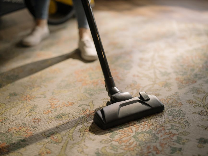 Close-up of a vacuum cleaner attachment being used on a patterned, woven area rug in a well-lit room, with visible parts of a person’s legs and shoes in the background. The rug features intricate floral motifs in muted tones, and appears clean and well-maintained. The vacuum head is black and rectangular, with a switch on top, and is positioned to effectively clean the surface. The scene emphasizes thorough surface cleaning and deep cleaning techniques suitable for residential spaces, highlighting attention to hygiene and maintenance. Carpet Cleaning SE10 offers same day emergency carpet cleaning in SE10 Greenwich, ensuring prompt sanitisation and domestic cleaning services to maintain a hygienic living environment.